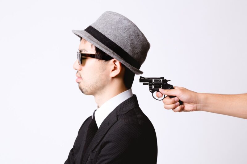 サングラスと帽子を身につけたスーツ姿の男性が背後から銃を突きつけられる画像