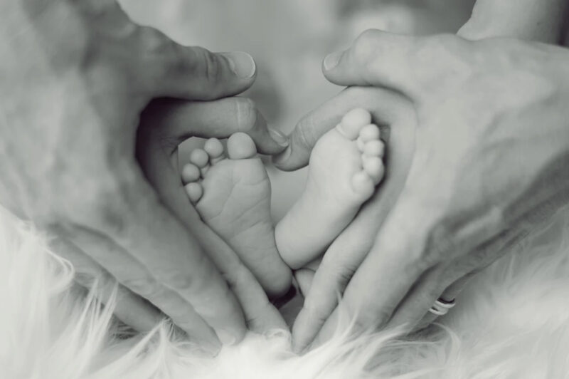 赤ちゃんの足で女性の手がハートを象り、その上を男性の手が同じくハートを象って覆っている画像