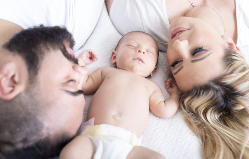 髭の男性と金髪の女性に囲まれて赤ちゃんが眠る様子の写真