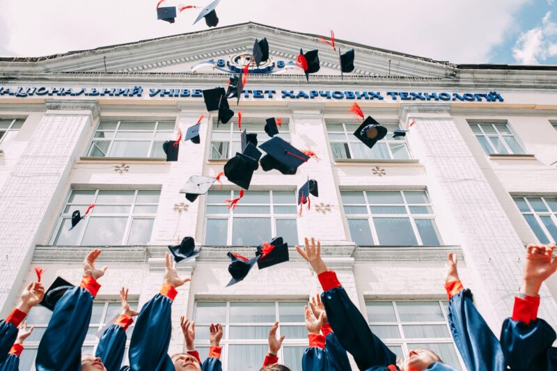 大学卒業生たちが白い校舎の前で、帽子を飛ばしている様子の写真